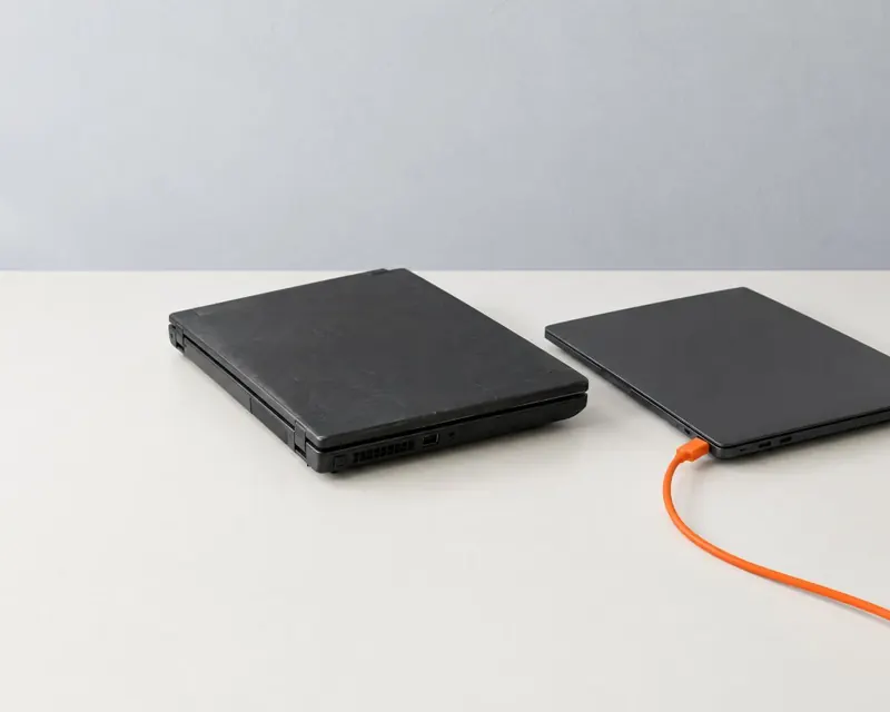 An older, scuffed laptop beside a newer one on a bench, the newer one plugged in with an orange cable.