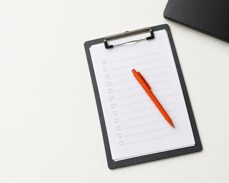 A clipboard with a blank checklist and an orange pen, beside a closed laptop.